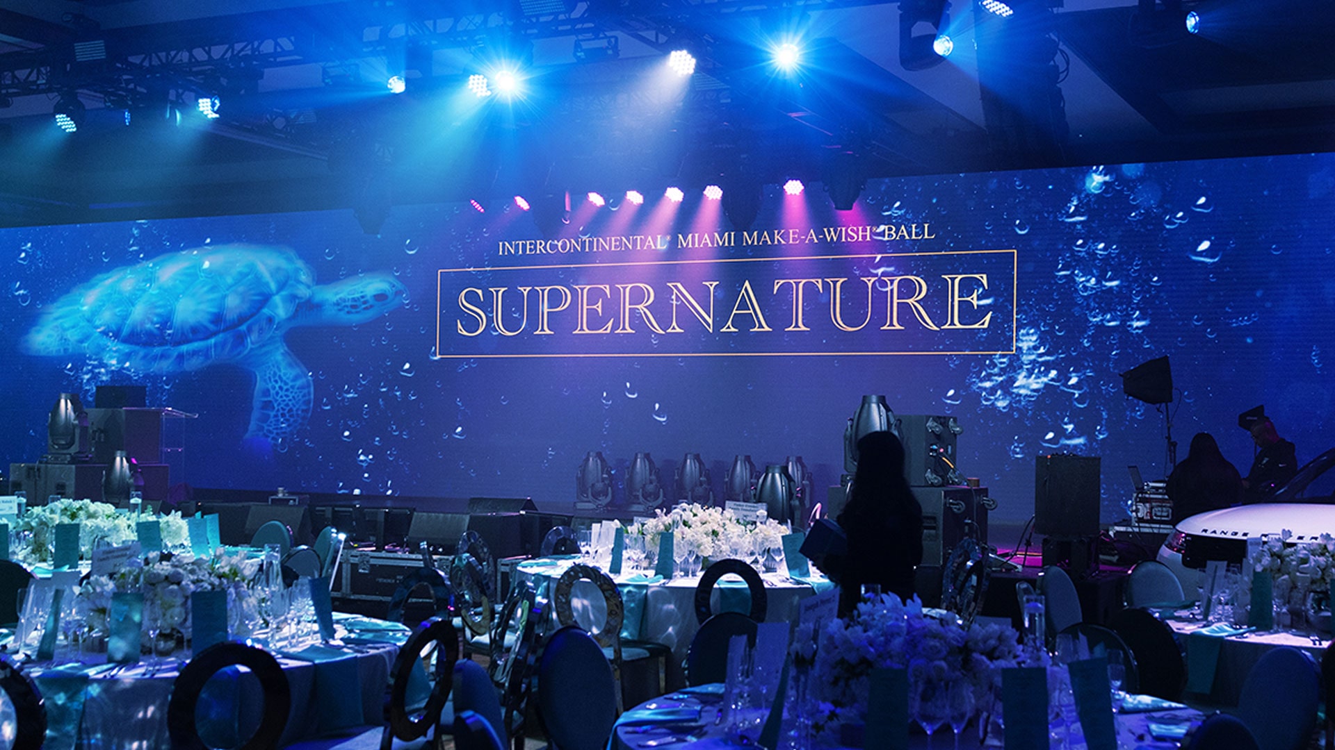 A decorated ballroom with tables set for an event. The stage backdrop displays a swimming sea turtle and the text “InterContinental Miami Make-A-Wish Ball: Supernature” under dramatic blue lighting.