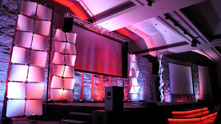 การจัดเวทีสมัยใหม่พร้อมจอโปรเจ็กเตอร์ขนาดใหญ่ แผงไฟรูปทรงเรขาคณิต ลำโพง และแสงไฟสีชมพูและม่วงที่ดูน่าตื่นตาตื่นใจ พร้อมสำหรับการนำเสนอหรือกิจกรรมในสถานที่จัดงานในร่มที่กว้างขวาง.