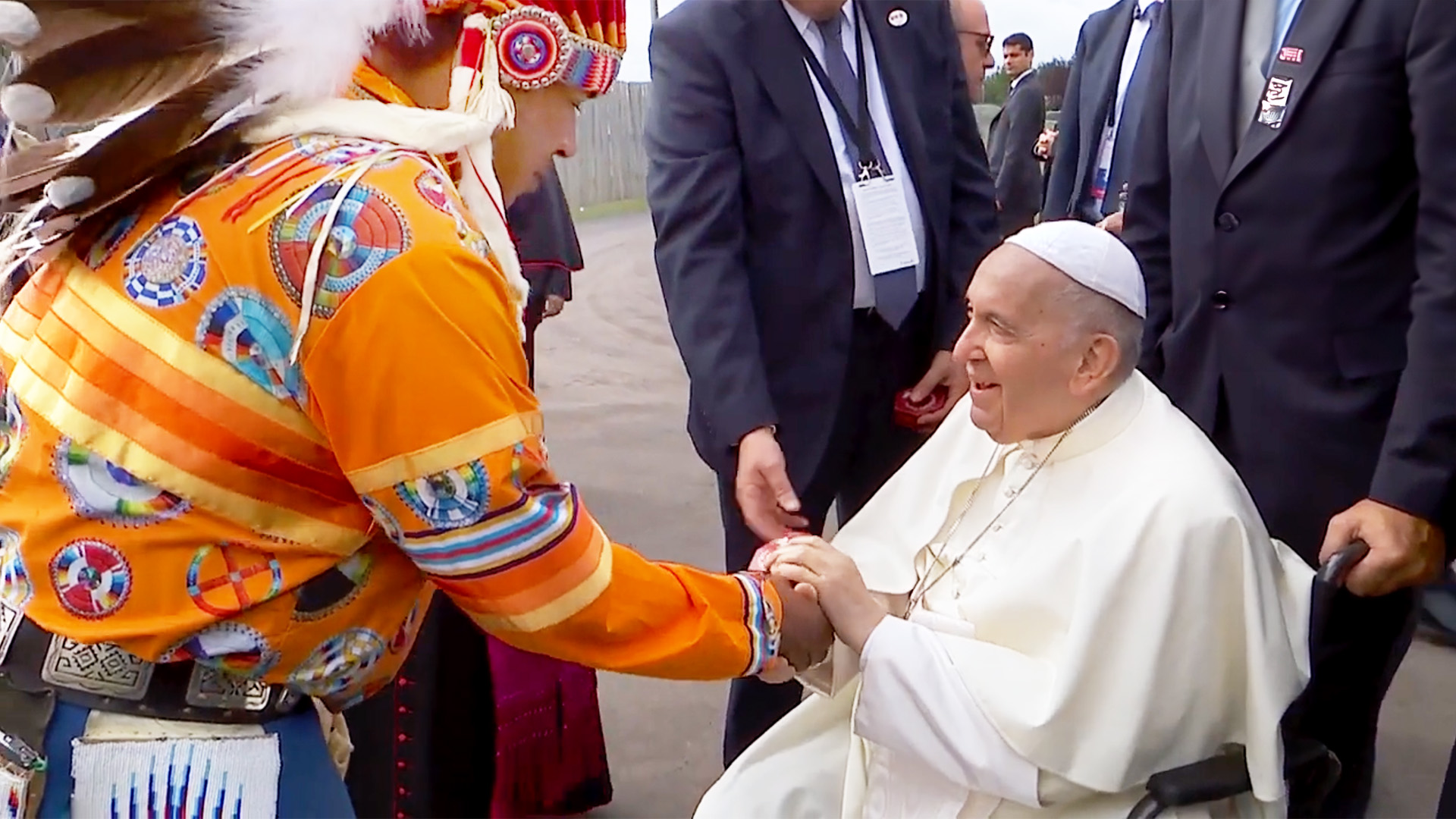 บุคคลในชุดประจำเผ่าพื้นเมืองอันสดใสกำลังจับมือกับผู้นำทางศาสนาสูงวัยที่ยิ้มแย้มในชุดคลุมสีขาวและหมวกทรงกลมสีดำ นั่งอยู่บนรถเข็น ล้อมรอบด้วยผู้คนในชุดสูท.