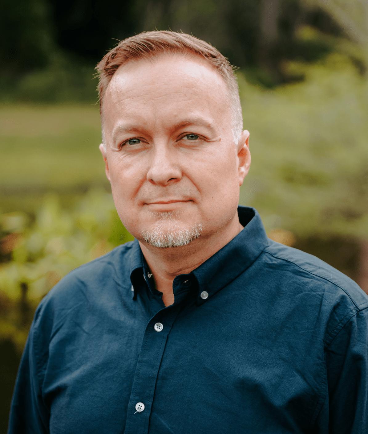 A man with short light brown hair and a trimmed goatee wears a dark blue button-up shirt, standing outdoors with greenery and blurred trees in the background.
