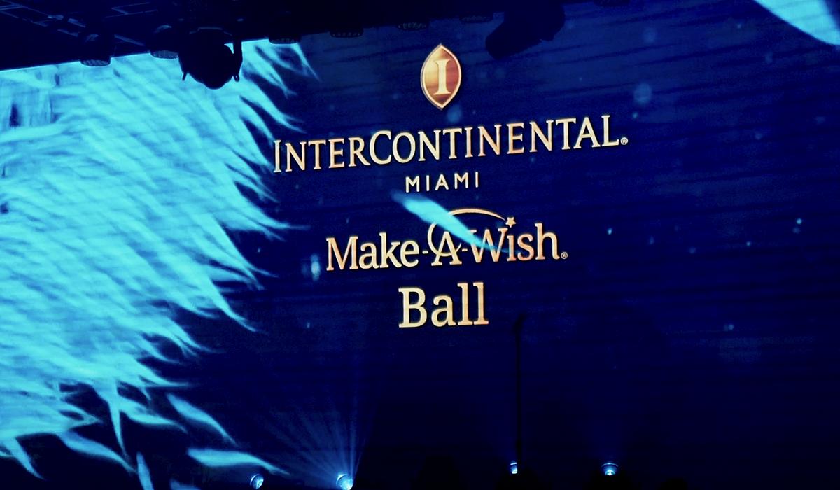 A large screen displays the text InterContinental Miami Make-A-Wish Ball with a blue, abstract background and spotlights shining upward.