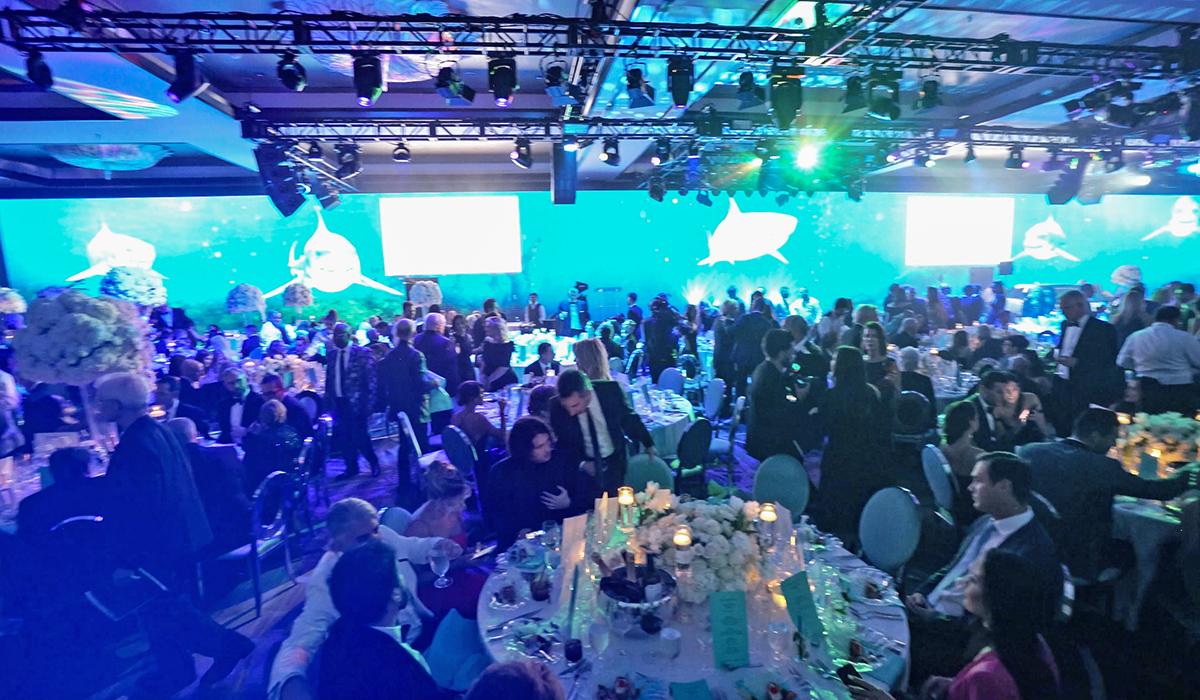 A large, elegant banquet hall filled with people seated at round tables, decorated with flowers and candles. The room is lit by colorful lighting and large screens with blue aquatic-themed visuals are displayed on the walls.