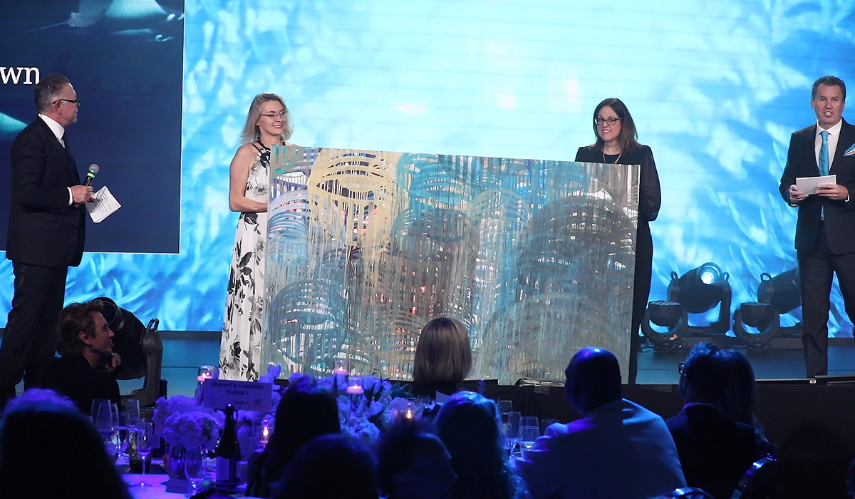 Four people stand on stage during an event, with two women holding a large abstract artwork. The background features blue lighting, and an audience is seated at tables in the foreground.