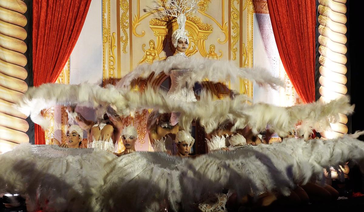 A group of performers in elaborate feathered costumes dances on stage with red curtains and ornate golden decorations, centered around one performer elevated above the others.