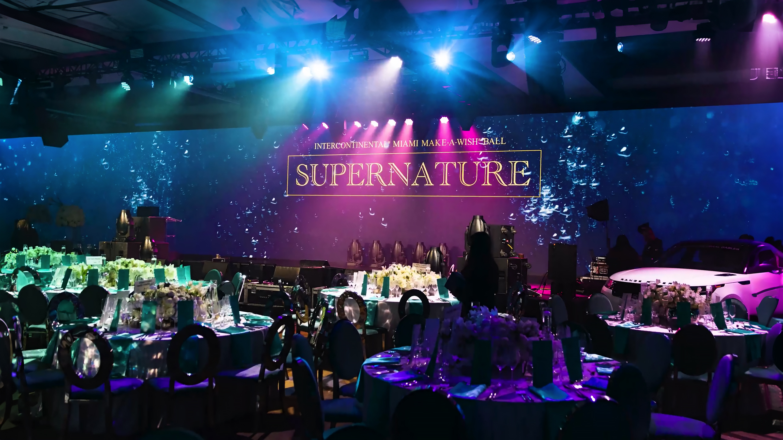 A large, elegant banquet hall with round tables set for an event, vibrant purple and blue lighting, and a large screen on stage displaying SUPERNATURE at the InterContinental Miami Make-A-Wish Ball.
