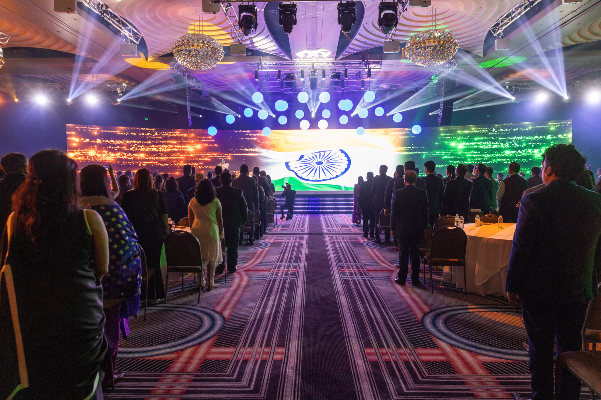 People in formal attire stand facing a stage with a large digital display of the Indian flag, in a grand, well-lit hall with chandeliers and patterned carpet, suggesting a formal ICICI Prudential Life event or ceremony.