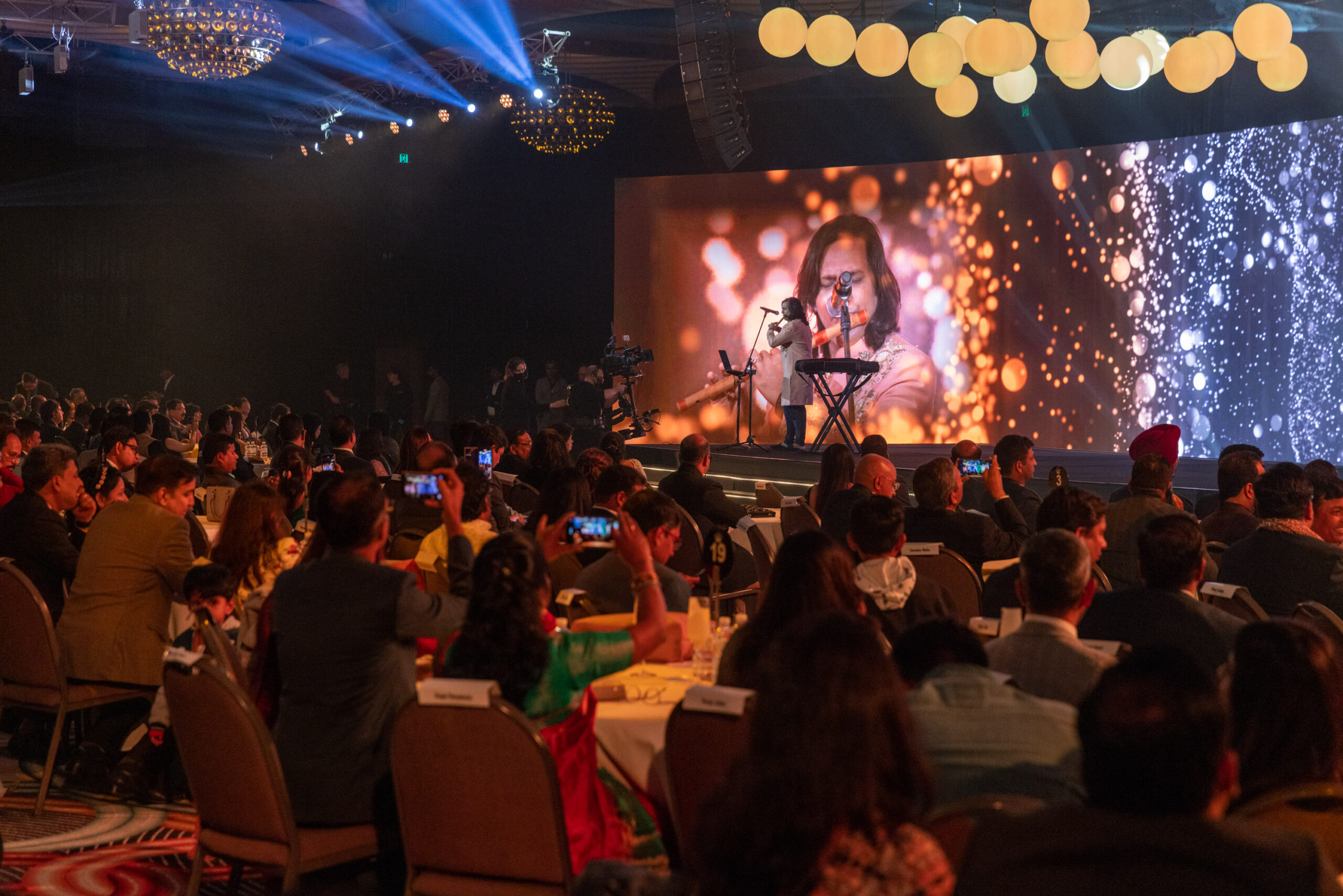 A musician performs on stage with a keyboard in front of a large audience, while a screen behind—sponsored by ICICI Prudential Life—shows a close-up of the performer with sparkling light effects. Audience members capture the moment on their phones.
