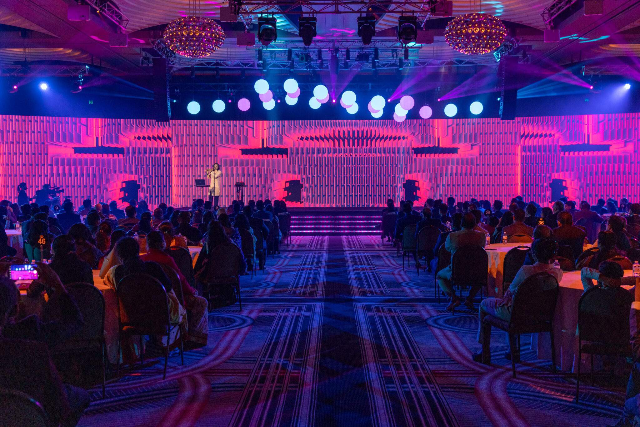 An audience sits at round tables in a large, dimly lit event hall, facing a brightly lit stage with purple and blue lighting effects where an ICICI Prudential Life presenter speaks at the podium.