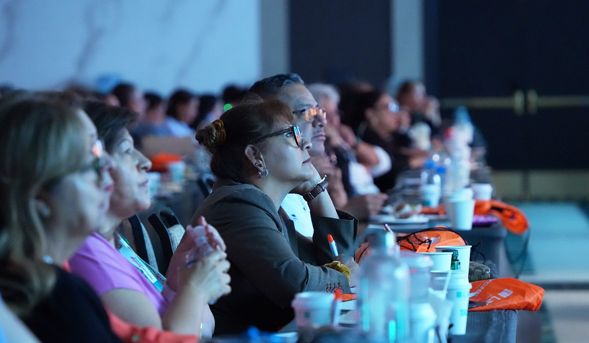Audience members, mostly women, sit at tables with drinks and notebooks in a dimly lit room, attentively watching a presentation or speaker off-camera.