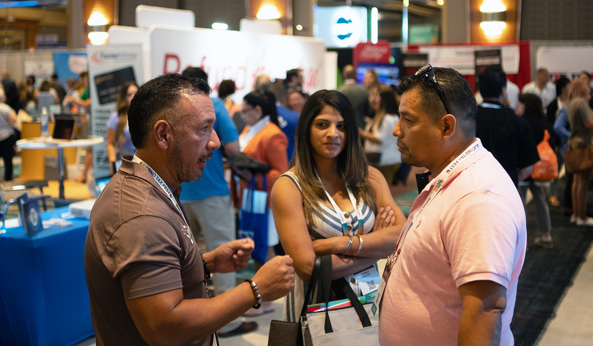 Three people stand and talk at a busy indoor convention or expo, wearing badges around their necks. Booths and other attendees are visible in the background.