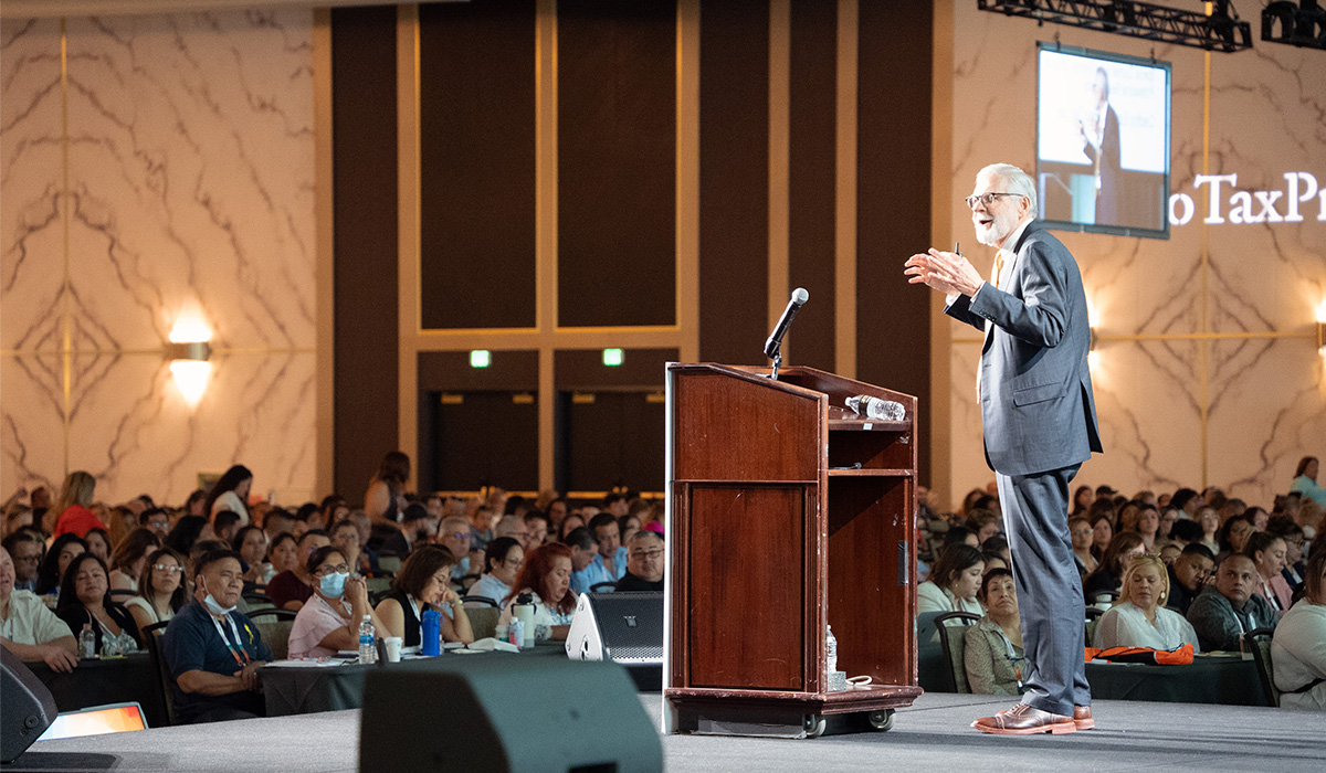 A man in a suit speaks at a podium on stage in front of a large audience at a conference; a screen and partial TaxP text are visible in the background.