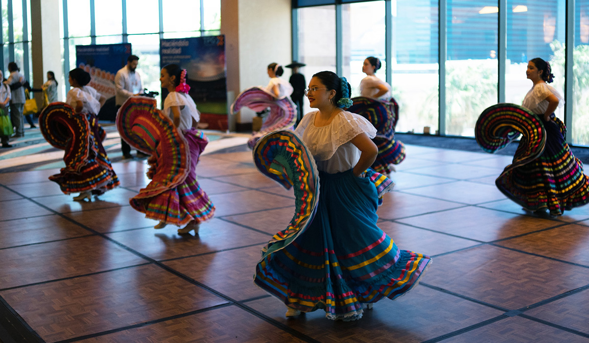 Women in traditional colorful dresses perform a folkloric dance indoors on a wooden dance floor, with natural light coming through large windows in the background.