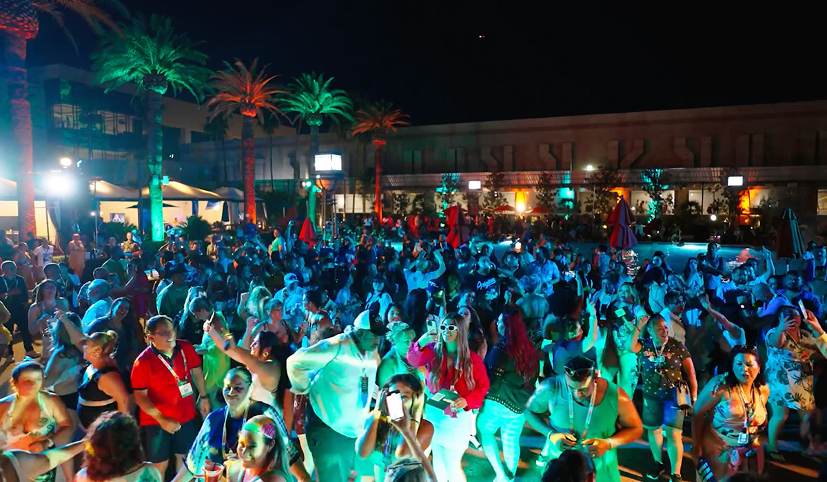 A large crowd of people enjoys a lively outdoor pool party at night, with colorful lights, palm trees, and vibrant decorations creating a festive atmosphere. Many are dancing, socializing, and taking photos.