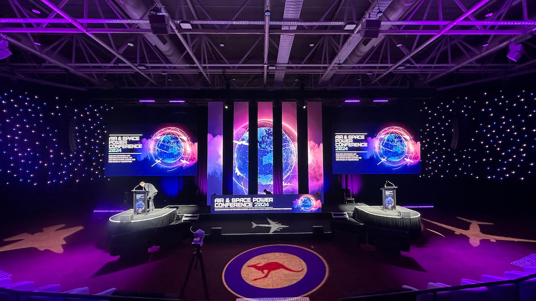 A stage setup for the Air & Space Power Conference 2024 features three large screens with a digital globe image, star-like lights on the walls, and silhouettes of aircraft on the floor. Two podiums and a kangaroo logo are visible.