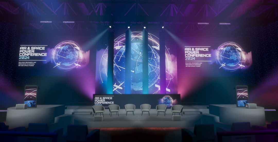A modern conference stage setup features large screens displaying Air & Space Power Conference 2024 with a digital globe graphic, illuminated by purple and blue lighting, and rows of empty chairs on stage.