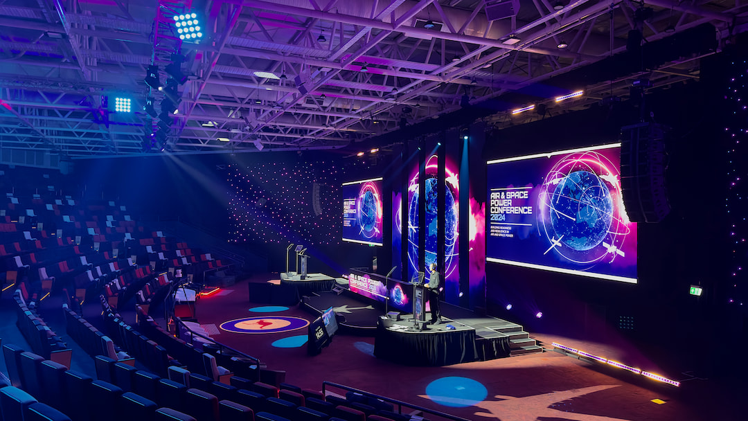 A modern conference hall with colorful lighting, large digital screens displaying RealSpace Pulse Conference 2023, a speaker on stage, and empty tiered seating surrounding the main floor.
