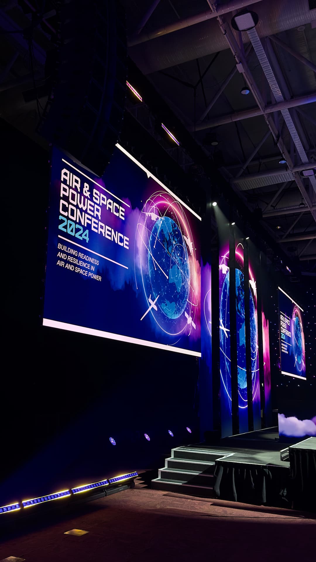 Large conference stage with illuminated screens displaying “Air & Space Power Conference 2024” and a digital globe graphic. The stage is set with steps and dramatic purple-blue lighting in a modern indoor venue.