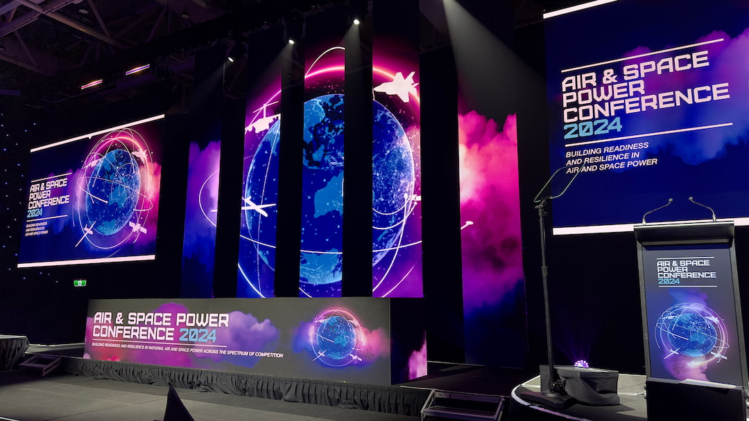 A stage set for the Air & Space Power Conference 2024 features large digital screens displaying a glowing Earth, conference title, and the theme “Building Readiness and Resilience in Air and Space Power.”.