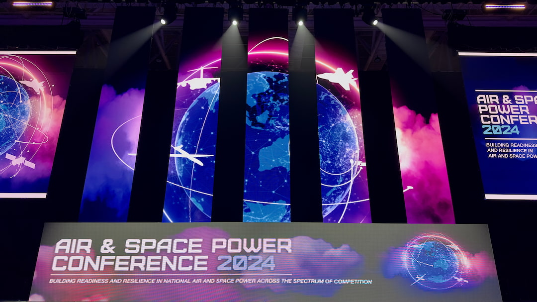 Colorful stage with large screens showing digital graphics of the Earth, airplanes, satellites, and text reading Air & Space Power Conference 2024 under bright spotlights.