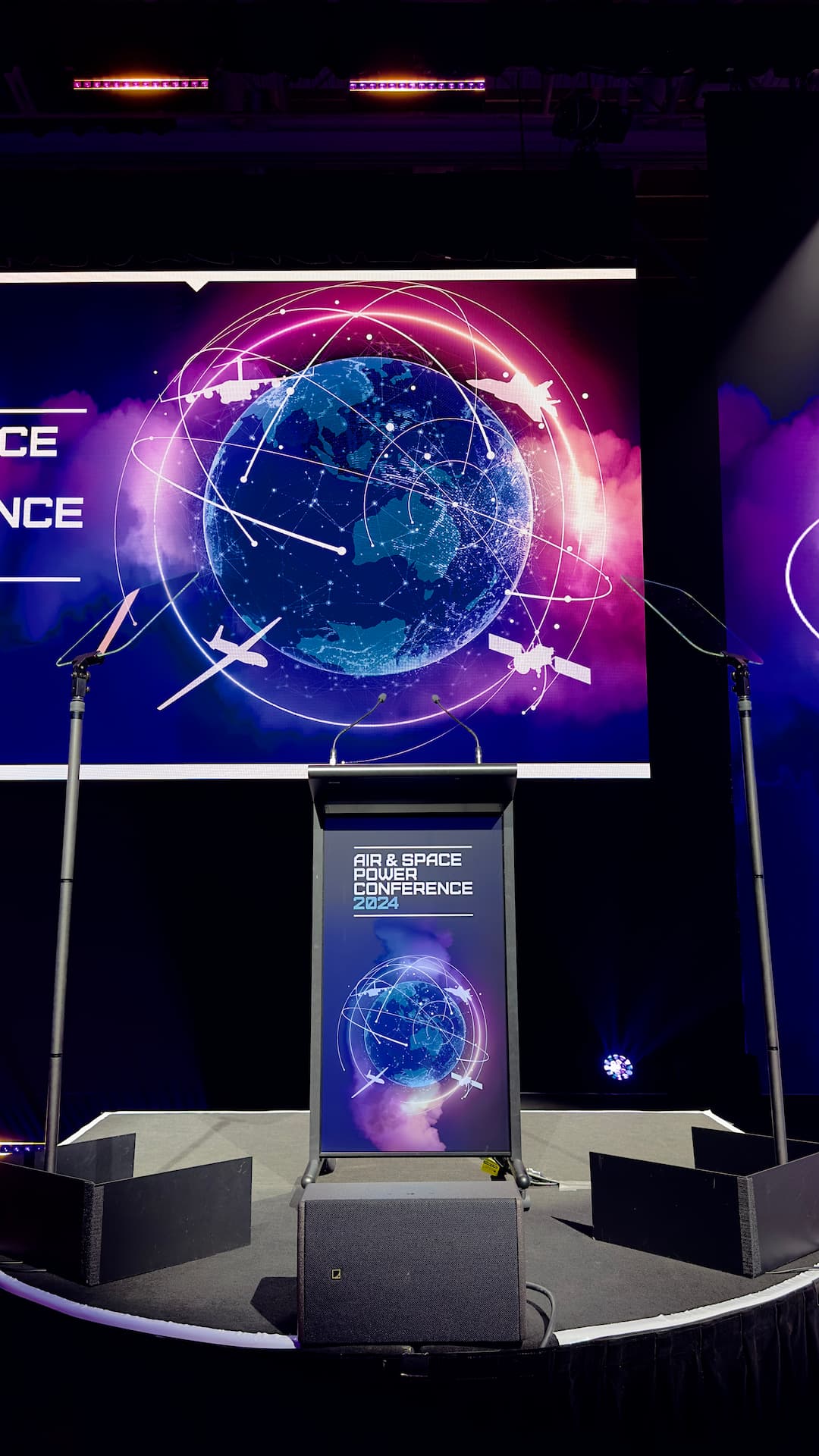 A stage with a podium set for the Air & Space Power Conference. Behind the podium, a large screen displays a digital illustration of Earth with satellites and aircraft orbiting, connected by glowing lines.