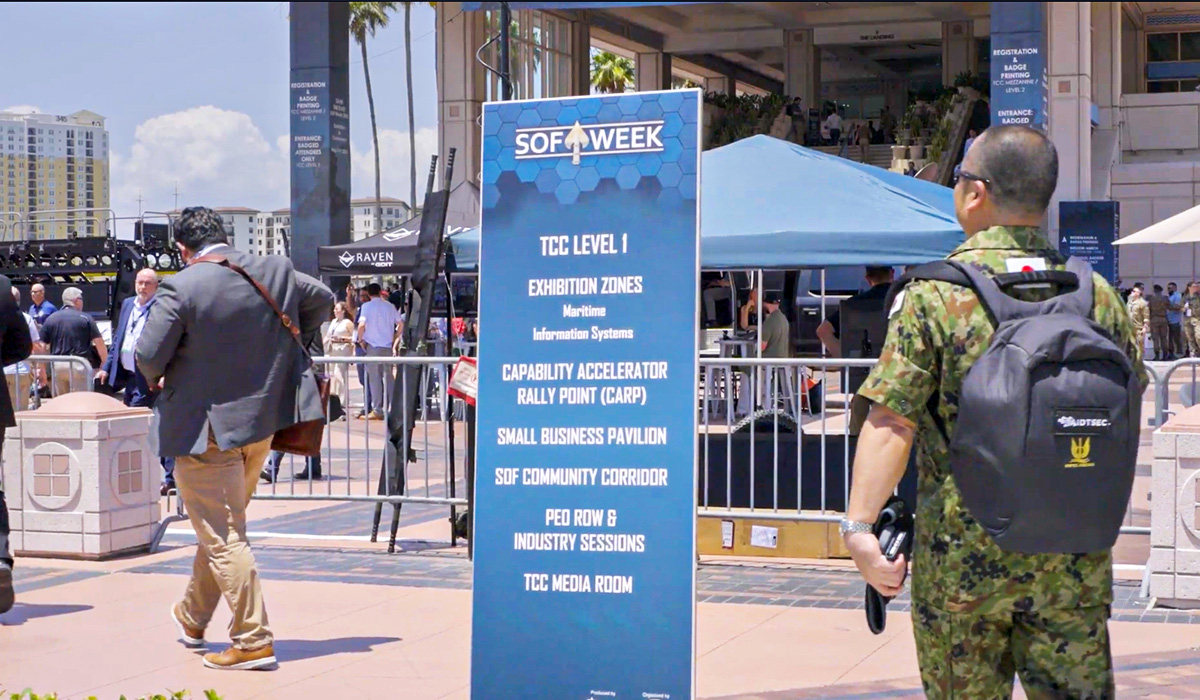 A large blue SOF Week event sign lists zones and activities as people, including one in camouflage, walk by an outdoor venue with tents and a crowd in the background on a sunny day.