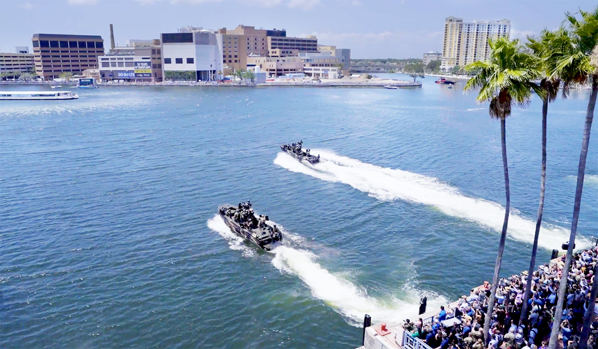 Two military boats speed across a harbor, leaving white trails in the water. A crowd watches from the waterfront, and modern buildings line the opposite shore under a clear, sunny sky with palm trees in the foreground.