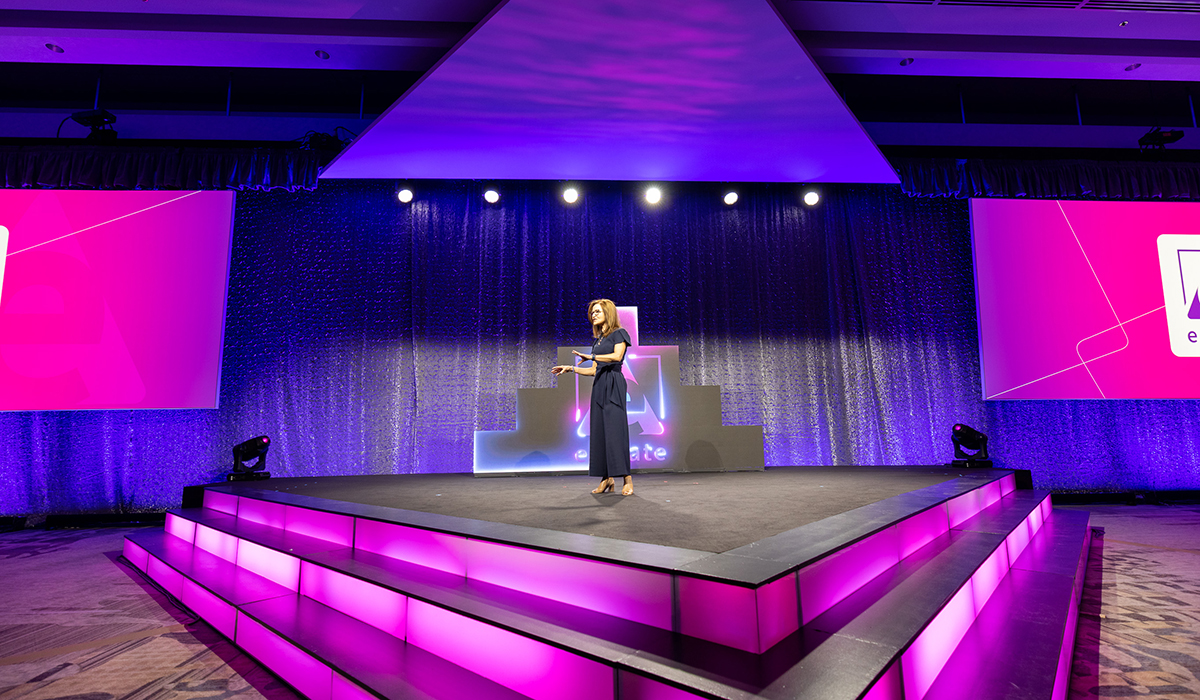 A woman stands on a brightly lit triangular stage with pink and purple lighting, speaking into a microphone. Two large screens with graphics flank the stage in a modern indoor event setting.