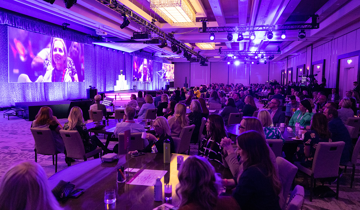 A large audience sits at round tables in a conference room, facing a stage with speakers and large projection screens displaying a woman speaking. The room is illuminated with purple lighting.