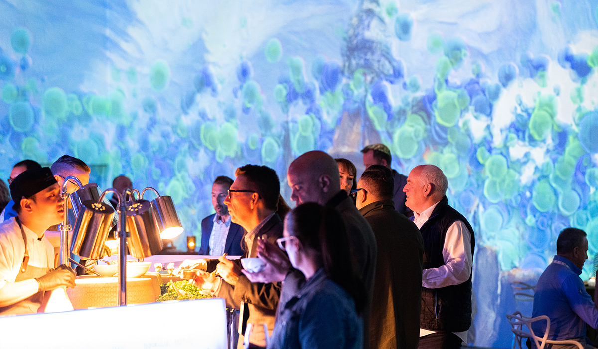 A group of people gather at a food station in a dimly lit room with vibrant blue and green abstract artwork projected on the walls. A chef serves food while guests converse and sample dishes.