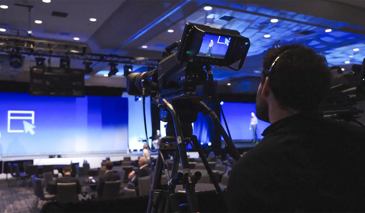 A cameraman films a speaker on stage at a conference in a large, modern auditorium with blue lighting and an audience seated in front of the stage.