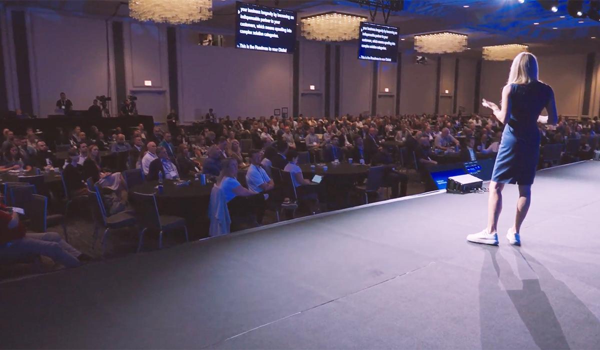 A woman stands on stage addressing a large seated audience in a conference hall. Two large screens display text above the crowd, and decorative chandeliers hang from the ceiling.