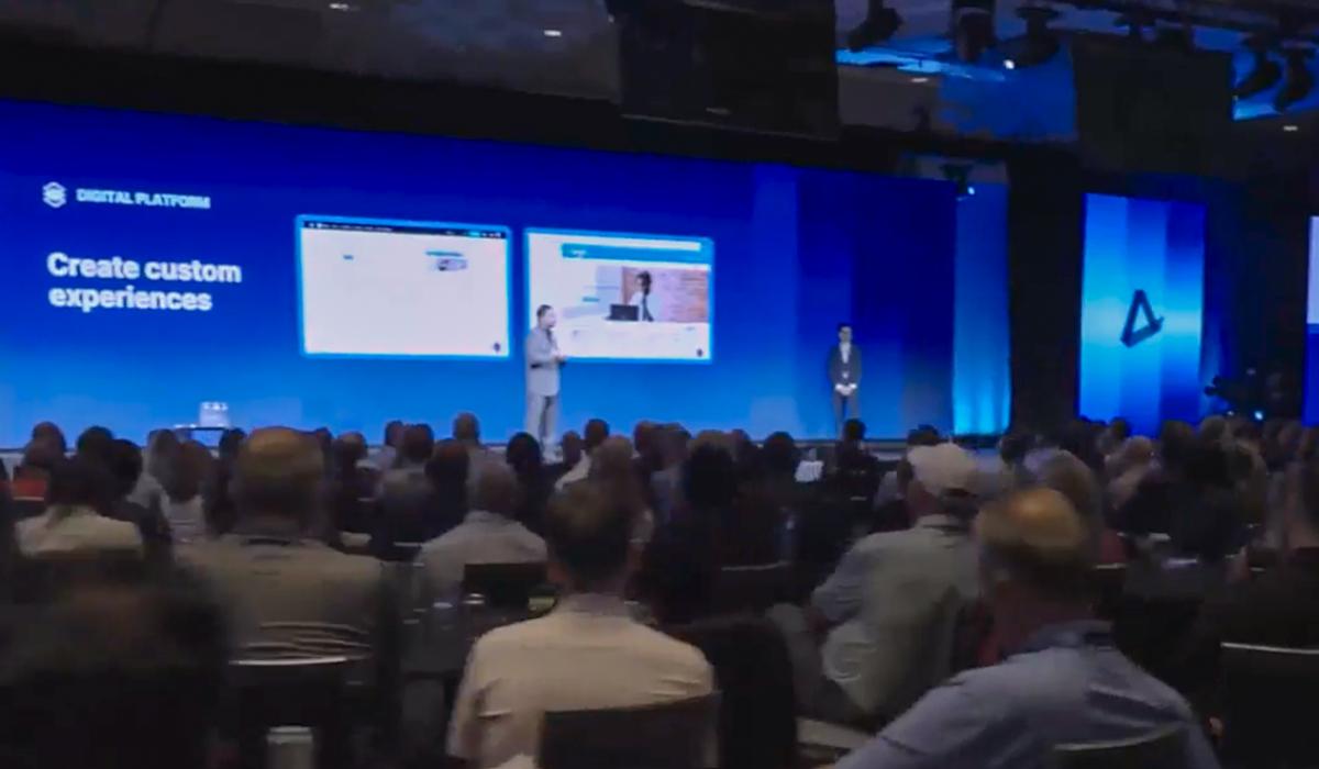 A large audience watches two presenters on stage at a conference. Behind them, screens display digital platform content and the words Create custom experiences on a blue background.