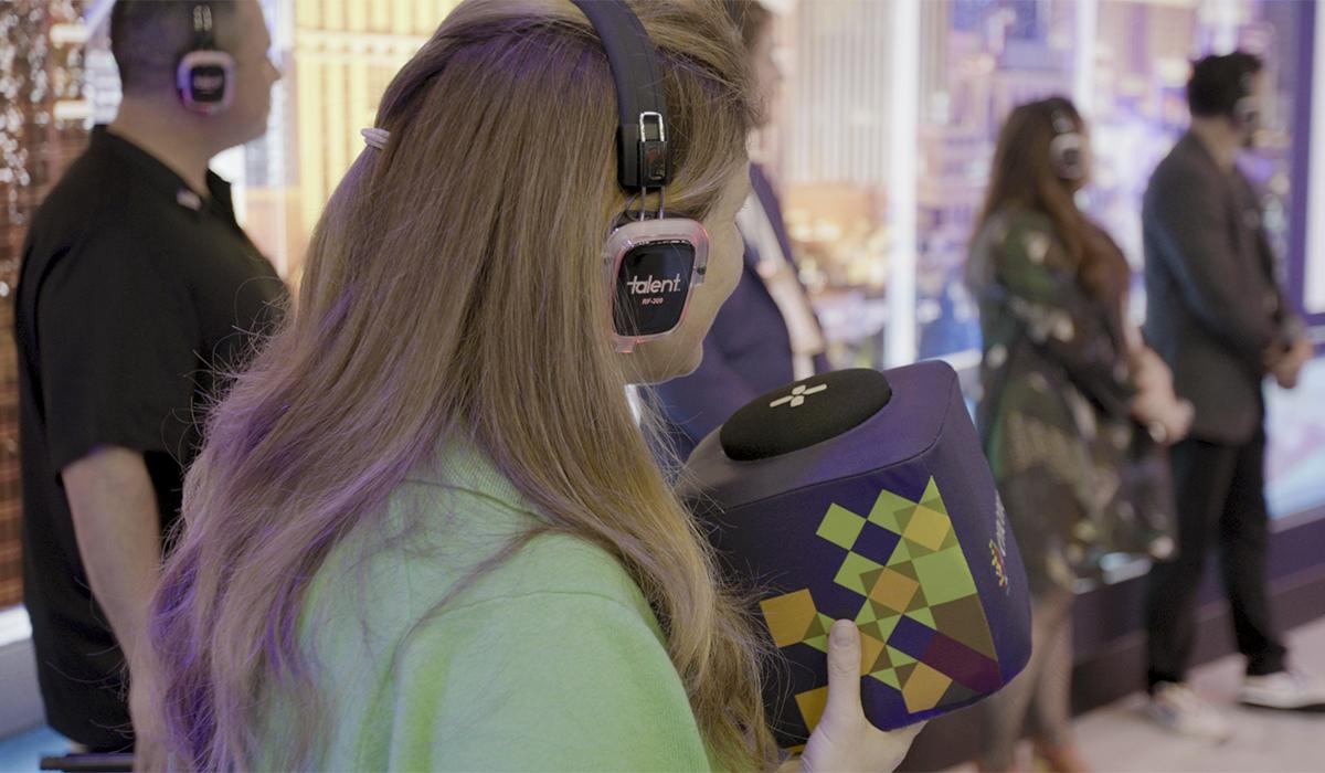 A person wearing headphones holds a colorful, cube-shaped object with a geometric pattern. Several people, also wearing headphones, stand in a line in the background inside a modern, brightly lit space.