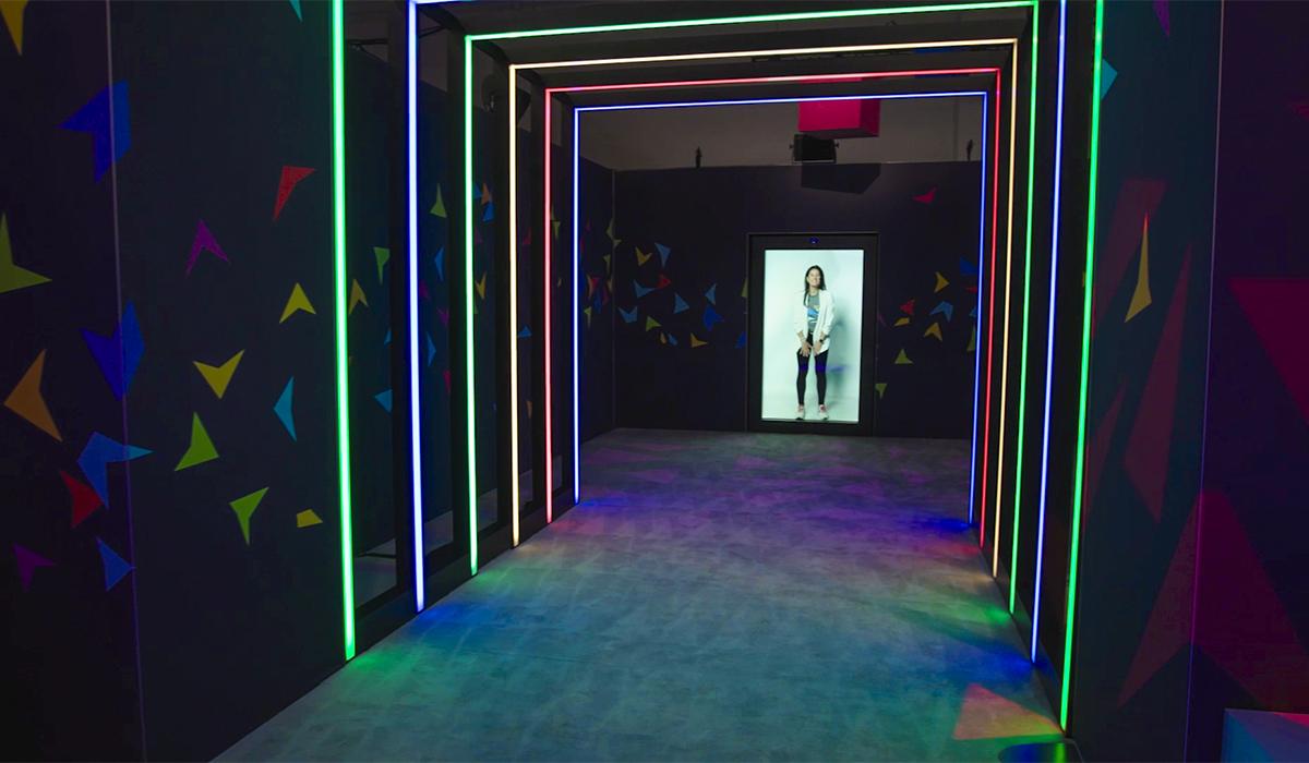 A woman stands at the end of a hallway lined with colorful, glowing LED light strips and geometric wall decorations, creating a futuristic and vibrant atmosphere.