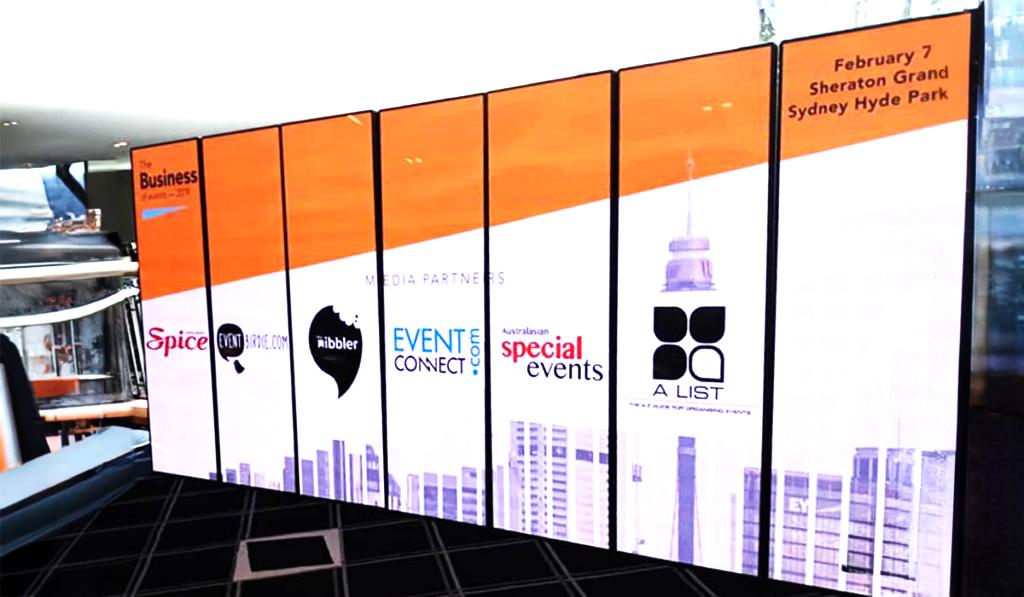 A large digital display shows event sponsors and details for a business event at Sheraton Grand Sydney Hyde Park on February 7. Logos for various media partners are visible on an orange and white background.