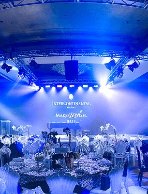 Elegant ballroom set for the InterContinental Miami Make-A-Wish Ball, with round tables, floral centerpieces, decorative chairs, and blue stage lighting illuminating the event logo on a screen.
