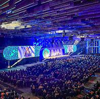A large audience sits in a spacious, modern auditorium with blue lighting, watching a presentation on a wide stage with multiple large screens displaying graphics and speakers.