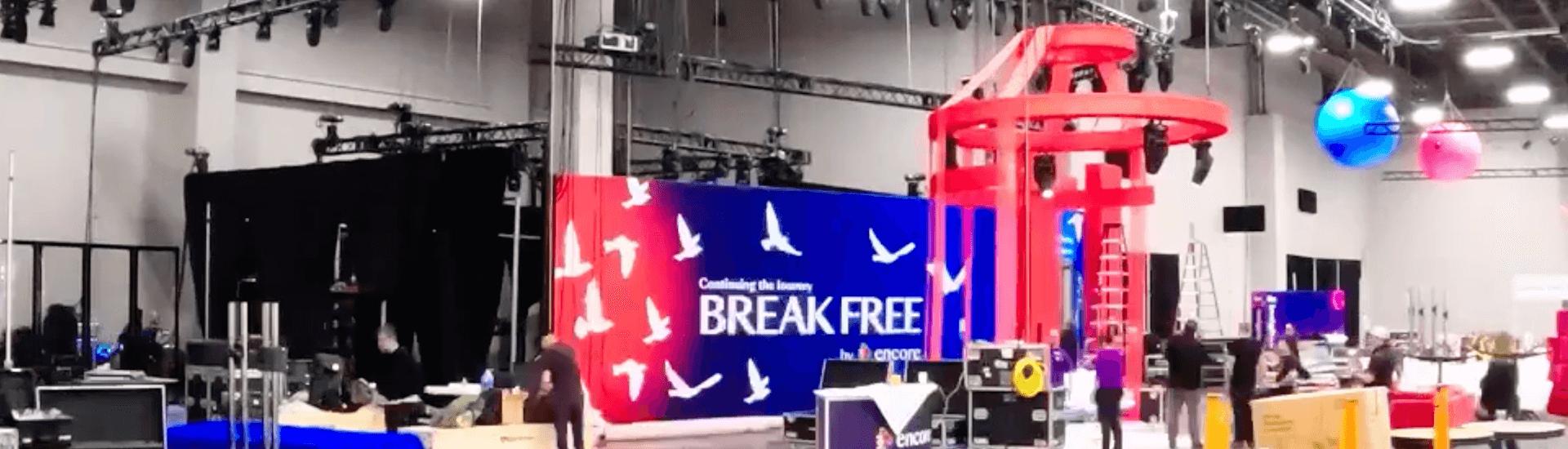 A large indoor event space is being set up for Break Free: IMEX America 2023 | Encore®, featuring a blue and red stage backdrop with white birds and the words “BREAK FREE.” Several people work as colorful decorations hang above.