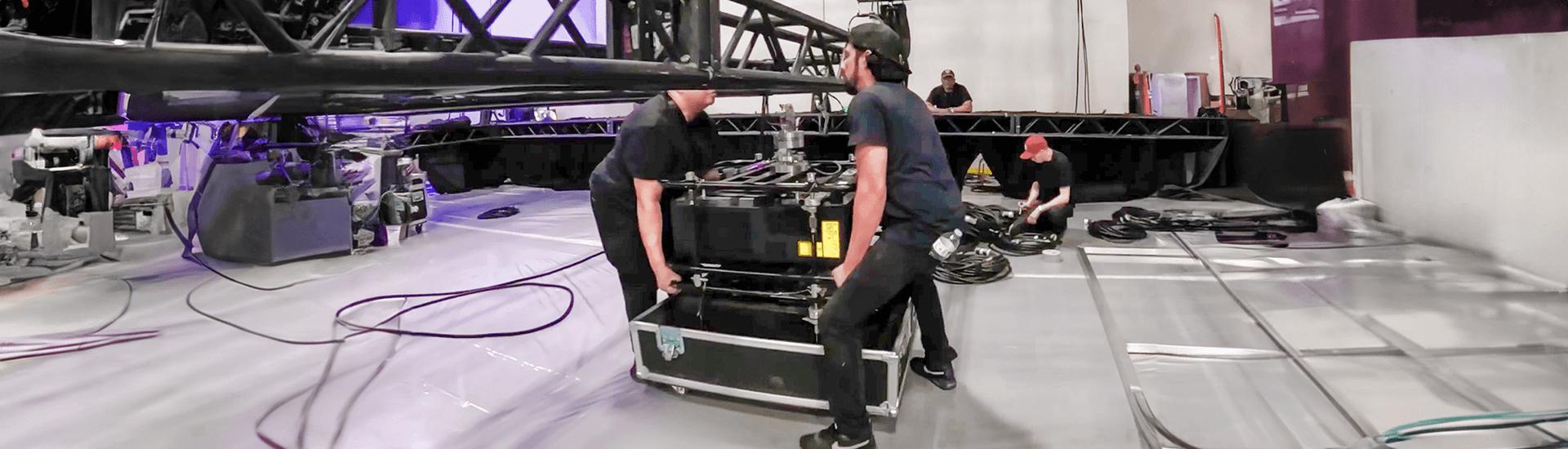 Several crew members set up stage equipment in a large indoor space for Break Free: IMEX America 2023 by Encore®, moving cables and gear cases under metal trusses on a glossy floor.