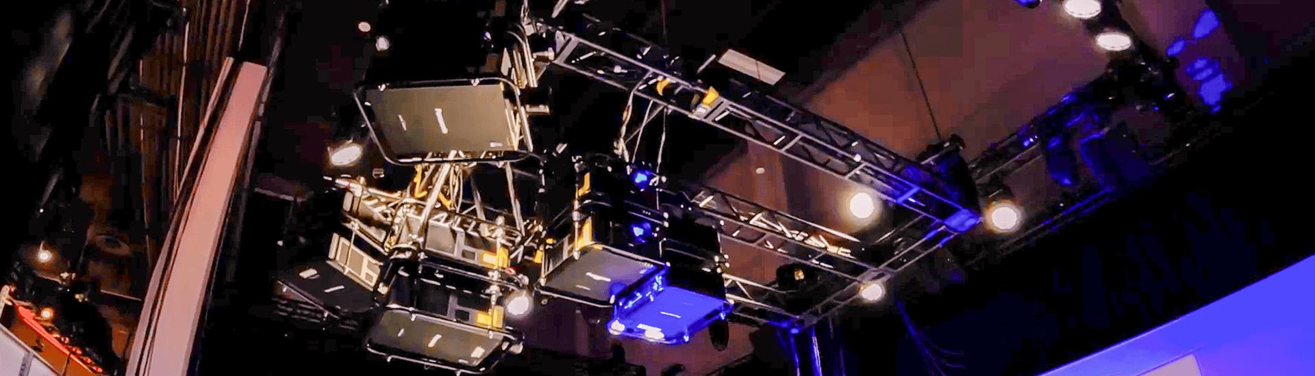 A lighting rig with multiple spotlights and stage lights, featured at Break Free: IMEX America 2023 by Encore®, is suspended from the ceiling, illuminating a theater or event space with a mix of purple and white lighting.