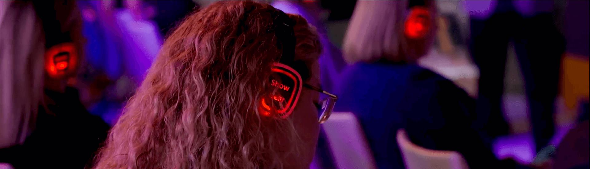 A person with curly hair wearing glowing red headphones at Break Free: IMEX America 2023 by Encore®, seated among others in a dimly lit room with purple lighting.