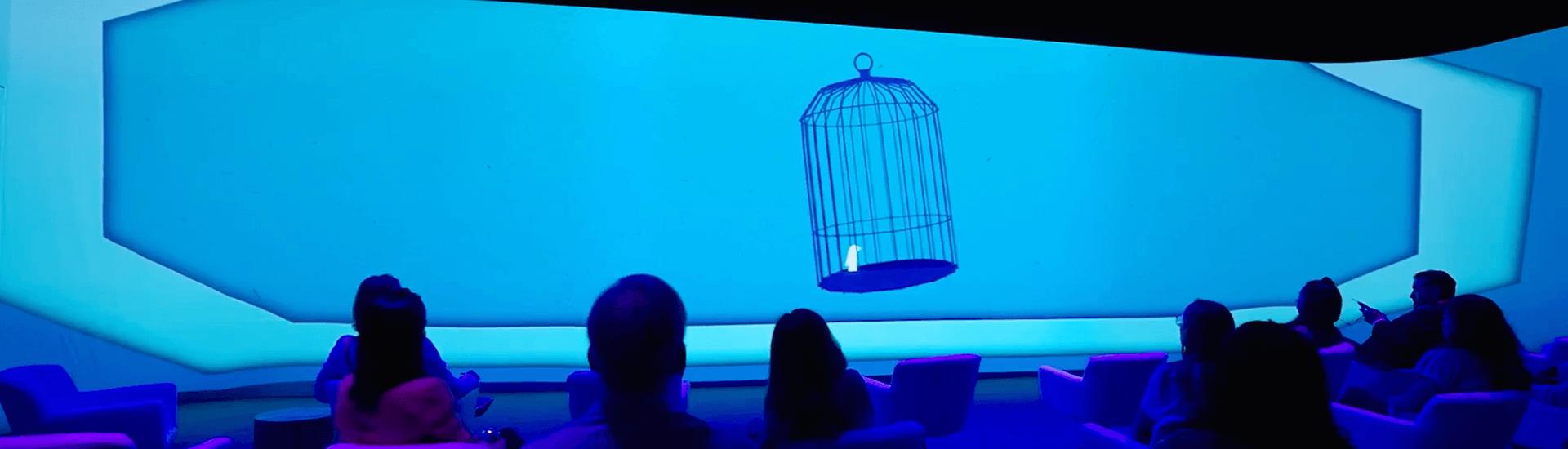 A group of people seated in a dark room at Break Free: IMEX America 2023 watch a large blue screen displaying a single empty birdcage with a swing, suspended in the center against a plain background—an experience by Encore(R).