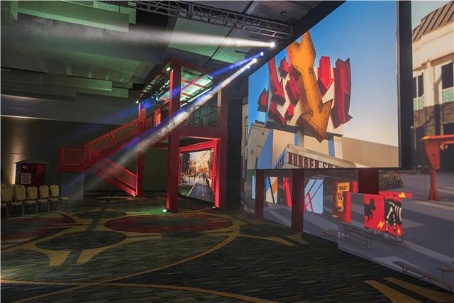 A large indoor event space features colorful abstract art projected on walls, rows of empty chairs, and a red staircase structure illuminated by stage lights. The floor is patterned with geometric designs.