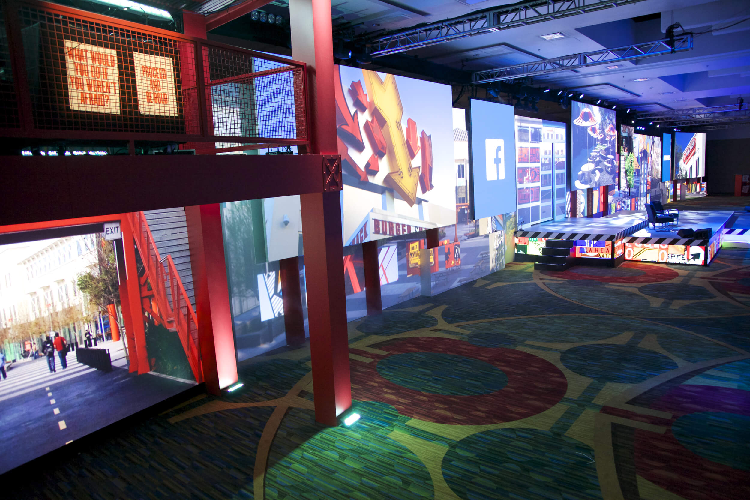 A large, modern indoor event space with colorful digital screens displaying artwork, cityscapes, and the Facebook logo. The carpet has geometric patterns, and the area is lit with vibrant, multicolored lighting.