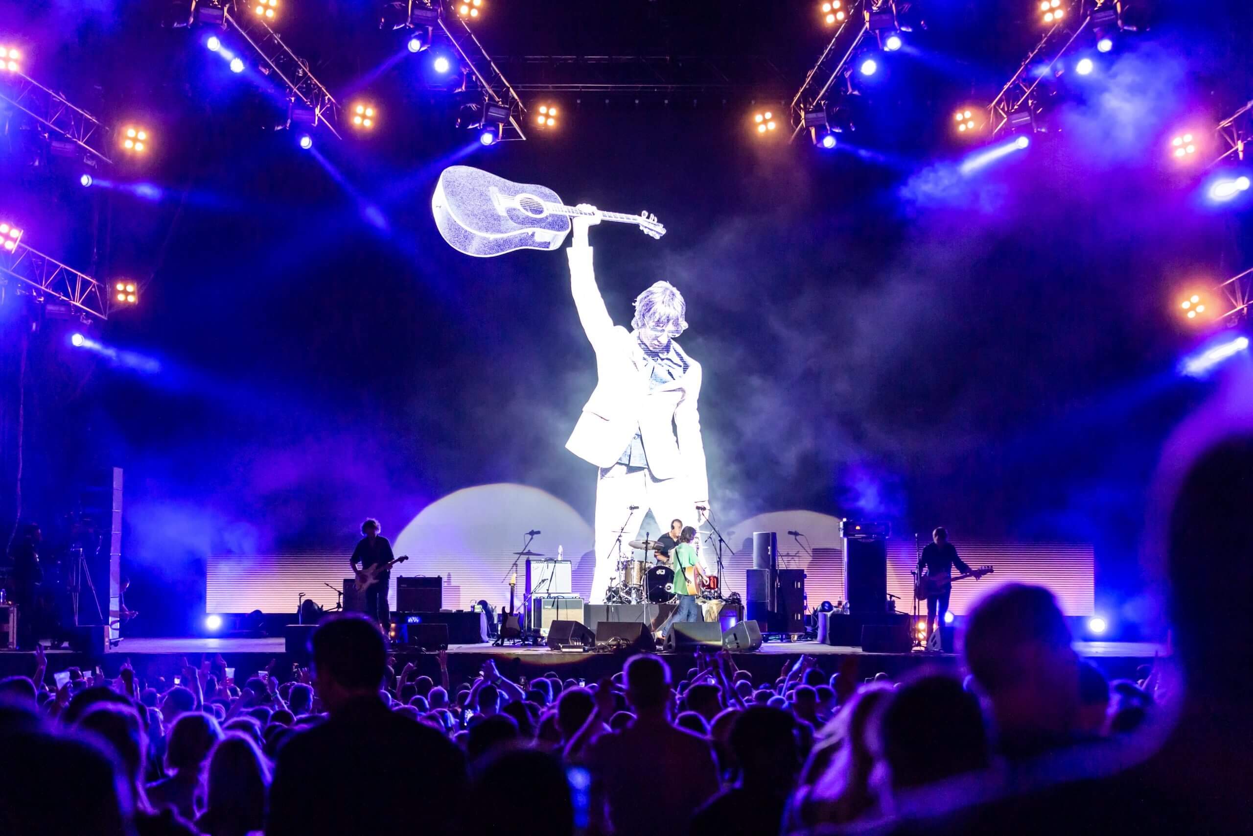 A large crowd watches a concert at night with bright stage lights and a giant screen displaying a black-and-white image of a person holding a guitar above their head. Several band members perform on the stage.