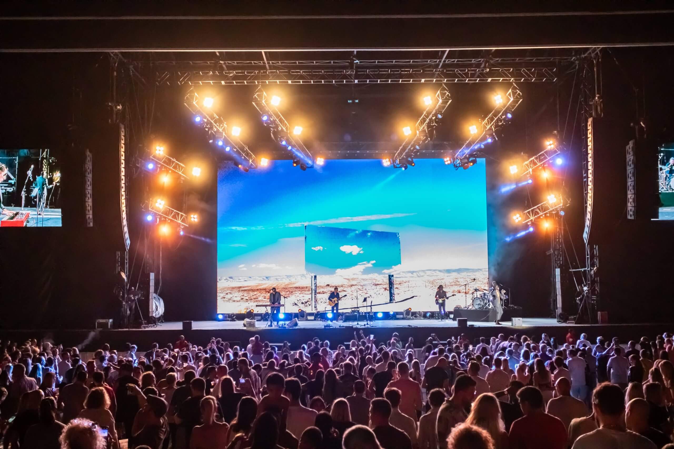 A large crowd watches a band perform on a brightly lit stage with a vivid desert landscape shown on a big screen behind them at an indoor concert venue.