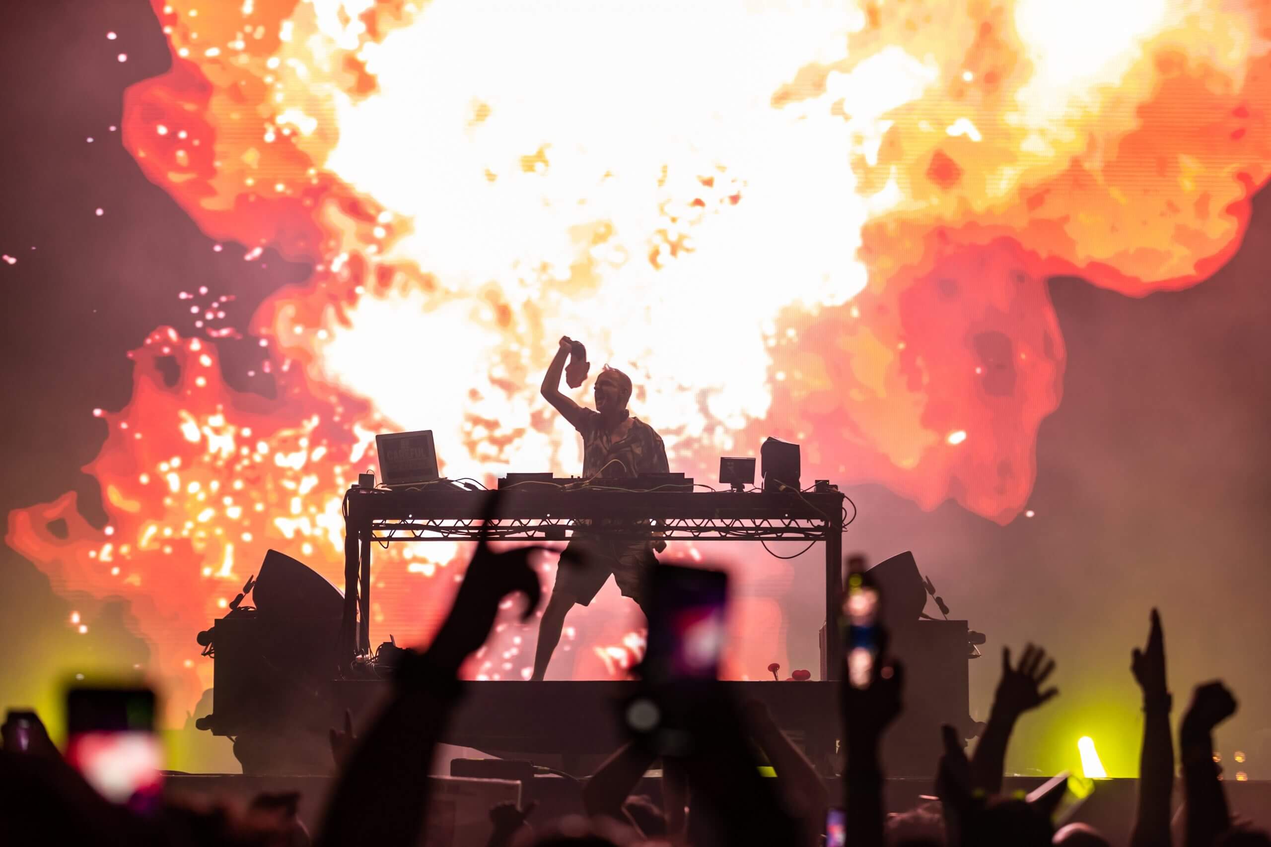 A DJ performs on stage in front of a large, fiery explosion backdrop while silhouetted fans in the crowd raise their hands and capture the moment with their phones.
