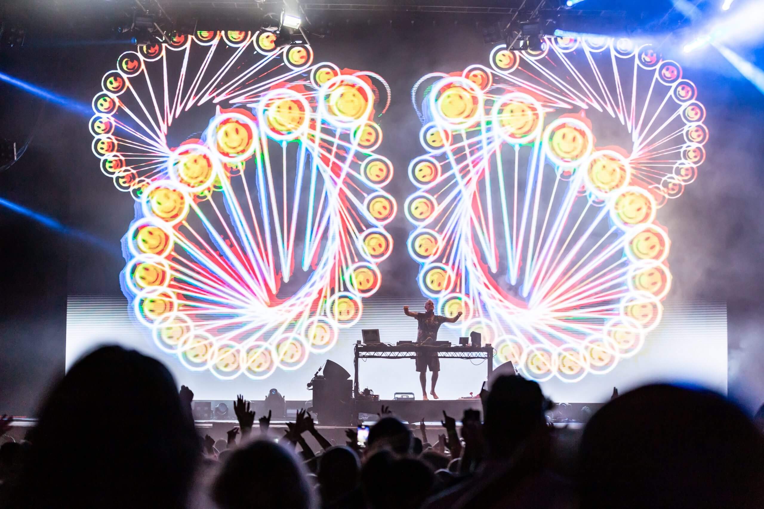 A DJ performs on stage with bright, colorful, spiral light patterns displayed behind them. The audience’s silhouettes can be seen enjoying the vibrant, energetic atmosphere of the concert.