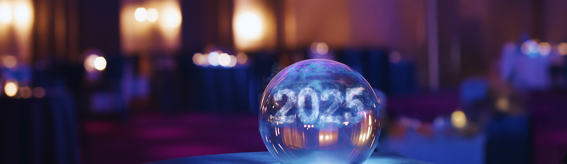 A glowing crystal ball on a table displays the number 2025 in sparkling text, with a softly lit, blurred background suggesting a dimly lit event or celebration.