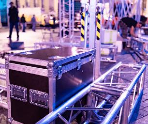 A large black equipment case sits on metal trusses at an outdoor event setup, with people and stage structures visible in the background under colorful lighting.